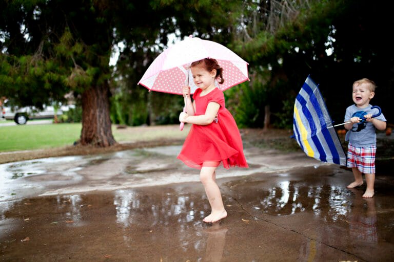 Young girl holding spotty pink umbrella in a puddle while a boy laughs