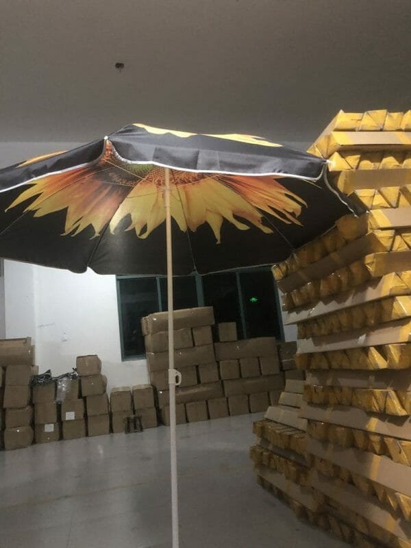 An 1.8M Polyester Black & Yellow Beach Umbrella with a sunflower design sitting in a warehouse.