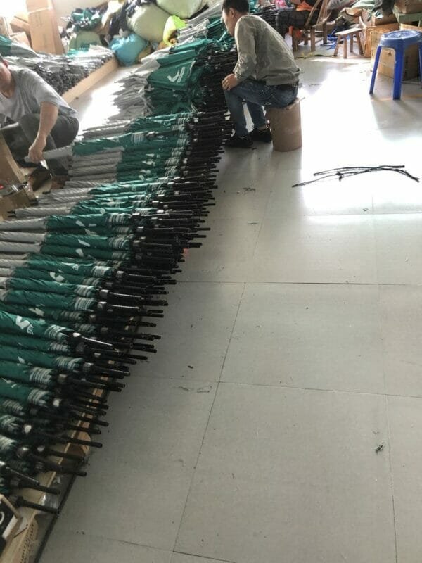 A group of people are working in a warehouse with a lot of 30inch Double Layer Golf Umbrella UV Coated umbrellas.