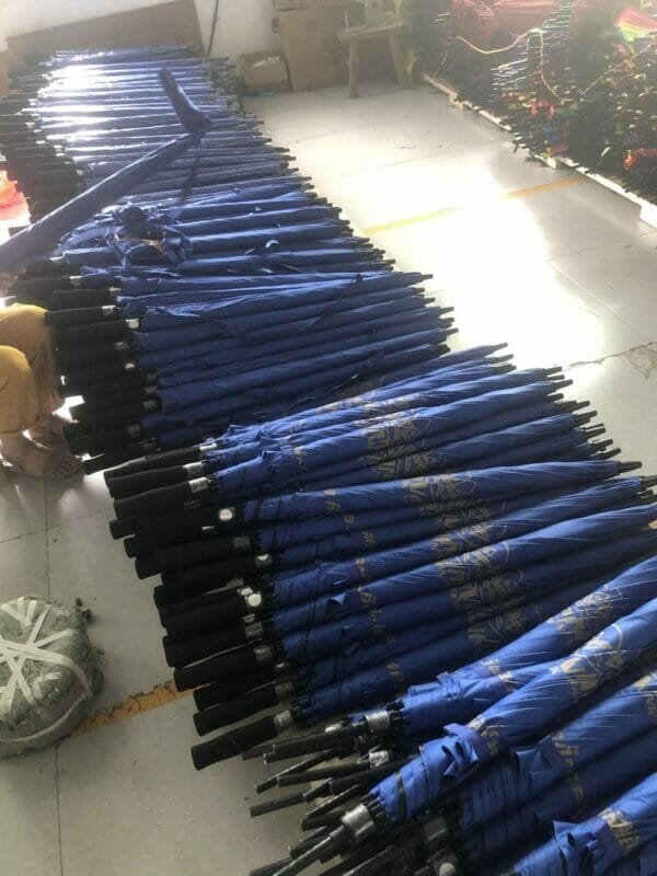 Many black single layer auto golf umbrellas are lined up in a warehouse.